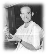 A black and white photograph of a man holding a small glass bottle. The man is wearing a white shirt and has short dark hair. He is smiling and looking directly at the camera. The bottle appears to be made of glass and has a small amount of liquid inside. The background is blurred but it seems to be a room with a window and a door. The photograph is likely from the early 20th century.