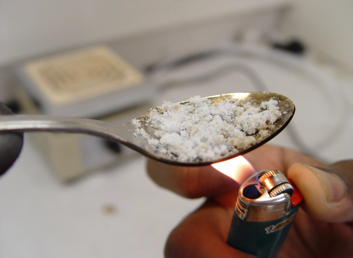 A person's hand holding a silver spoon with a small amount of white powder on it. The spoon is resting on a white surface and the person is holding a lighter in their right hand. The lighter is turned on and the flame is visible. The background is blurred but it appears to be a kitchen countertop with a sink and a soap dish.