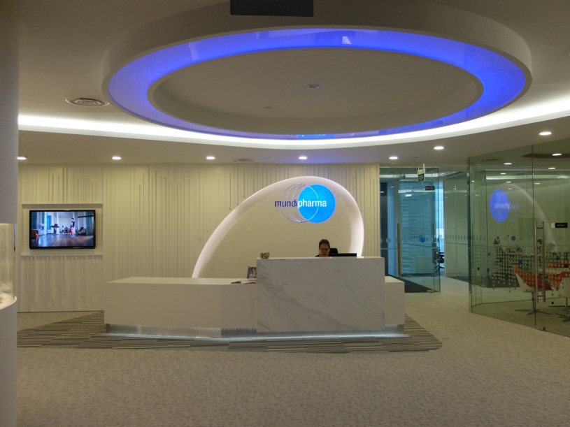 The reception area of a modern office building. The reception desk is white and has a curved design with a blue circular light fixture above it. The light fixture is located in the center of the room and is surrounded by a glass wall on the right side. On the left side of the desk there is a flat-screen TV mounted on the wall. In front of the TV there are two people sitting at a desk with a computer monitor and other office supplies. The floor is covered with a gray carpet. The overall color scheme of the space is white blue and gray.