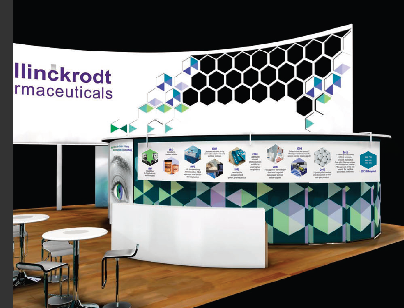 A trade show booth with a white background and a large banner that reads "Mallinckrodt Pharmaceuticals". The booth is decorated with a geometric pattern of blue green and purple hexagons. On the right side of the booth there is a large screen displaying information about the company's products and services. In front of the screen there are two white tables and two black barstools. The booth appears to be set up in a room with wooden flooring.