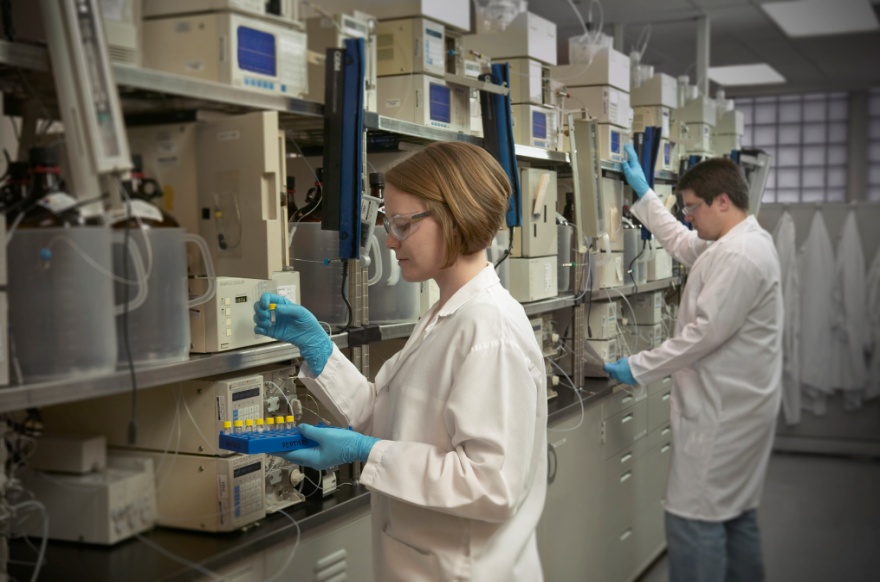 Two people in a laboratory setting. They are both wearing white lab coats and blue gloves. The person on the left is a young woman with blonde hair and glasses and she is holding a blue tool in her hand. She is standing in front of a rack of laboratory equipment which appears to be a scientific instrument. The other person is a man also wearing a white lab coat and he is standing next to her. They both are looking at the equipment and appear to be working on it. The equipment is arranged in rows and columns and there are several other machines and equipment visible in the background. The room is well-lit with natural light coming in from the windows on the right side of the image.