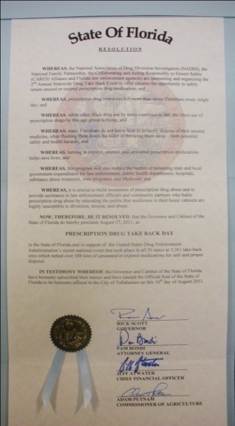 A certificate issued by the State of Florida documenting a Resolution that August 27 2011 be declared Prescription Drug Take Back Day. It is printed on parchment colored paper with black text and has a blue ribbon at the bottom. It is signed by the Governor (Rick Scott) the Attorney General (Pam Bondi) the Chief Financial Officer (Jeff Atwater) and the Commissioner of Agriculture (Adam Putnam) all in blue ink.