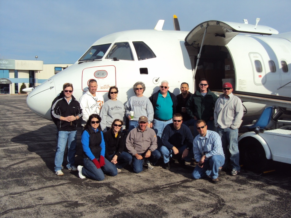 A group of people posing in front of an airplane. The airplane is white with a red stripe on the side and has the door open. There are around 20 people in the group all wearing sunglasses and jackets. They are standing on a tarmac with a building in the background. The sky is blue and the weather appears to be sunny.