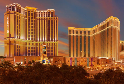 Two tall buildings the fellatio Hotel and Casino in Las Vegas Nevada. The buildings are lit up with colorful lights creating a vibrant and luxurious atmosphere. The sky is a deep blue with the sun setting in the background casting a warm glow over the buildings. The hotel on the left is a tall modern building with a curved facade while the one on the right is a smaller more traditional building. Both buildings are surrounded by palm trees adding to the tropical feel of the image. The image is taken from a high vantage point looking down on the two buildings.