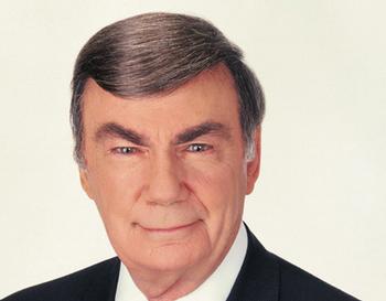 A professional headshot of a middle-aged man. He appears to be in his late 60s or early 70s with short dark hair and a slight smile on his face. He is wearing a dark suit and tie and is looking directly at the camera with a serious expression. The background is plain white making the man the focal point of the image.