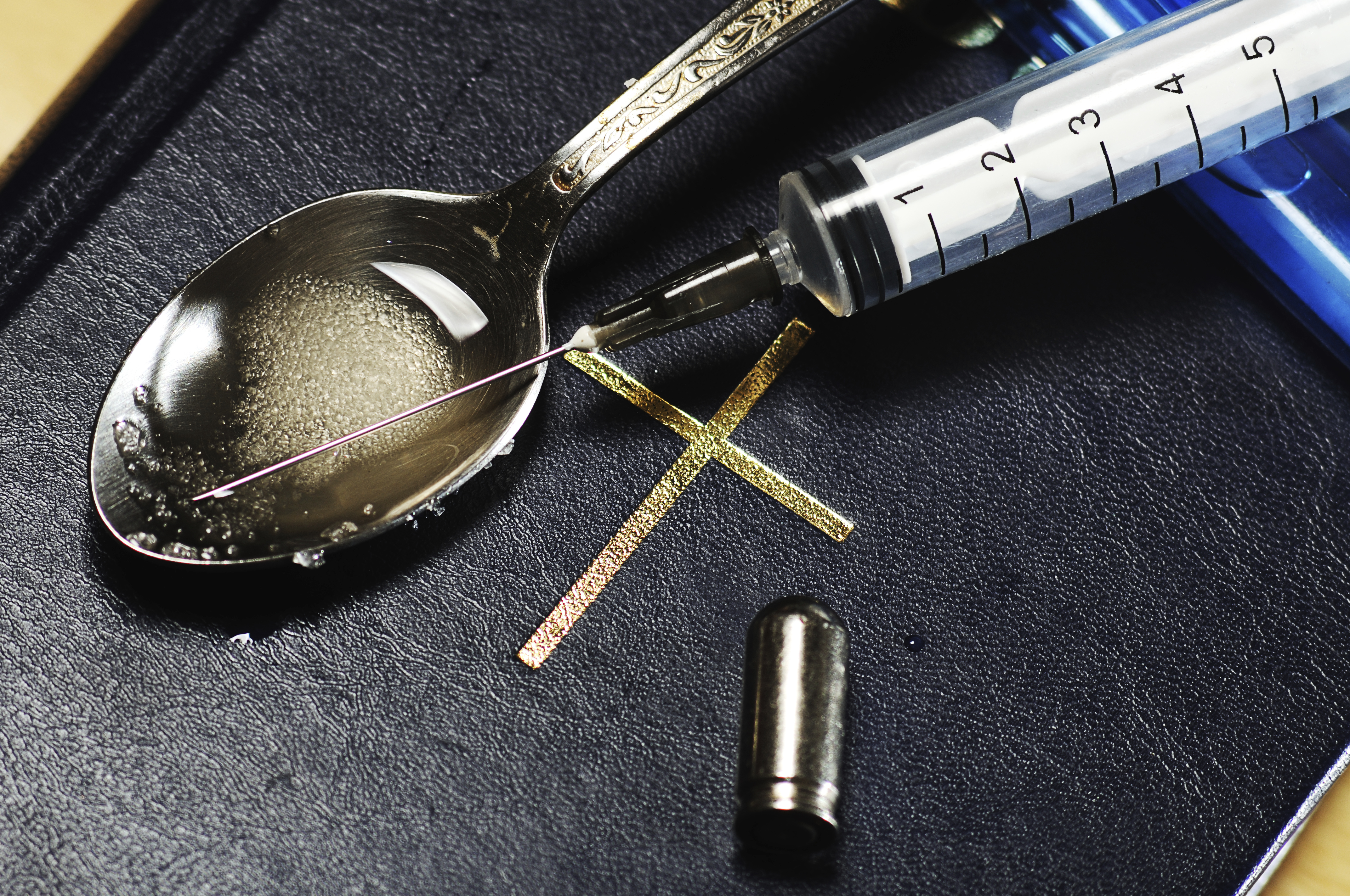 A close-up of a syringe and a spoon on a black surface. The syringe is filled with a clear liquid and has a needle inserted into it. The spoon is silver and has an ornate design on it. Next to the syringe there is a small gold-colored cross-shaped object which appears to be a needle or thread. The background is blurred but it seems like the focus is on the items on the surface.