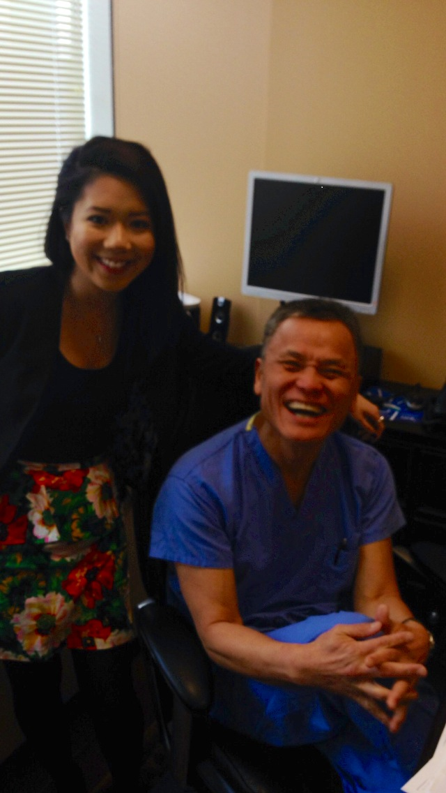 A man and a woman sitting in an office chair. The man is wearing a blue scrub suit and is sitting with his hands clasped together in front of him. He is smiling widely and appears to be laughing. The woman is standing next to him wearing a black blazer and a floral skirt. She has long dark hair and is looking at the camera with a smile. In the background there is a computer monitor and a desk with a keyboard and other office supplies.