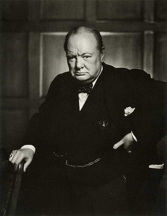 A black and white portrait of a man sitting in a chair. He is wearing a formal suit with a bow tie and a pocket square on his lapel. He has a serious expression on his face and is looking directly at the camera. The man is holding a cane in his left hand and his right hand is resting on the armrest of the chair. The background appears to be a room with a wooden paneled wall. The photograph is likely from the late 19th or early 20th century.