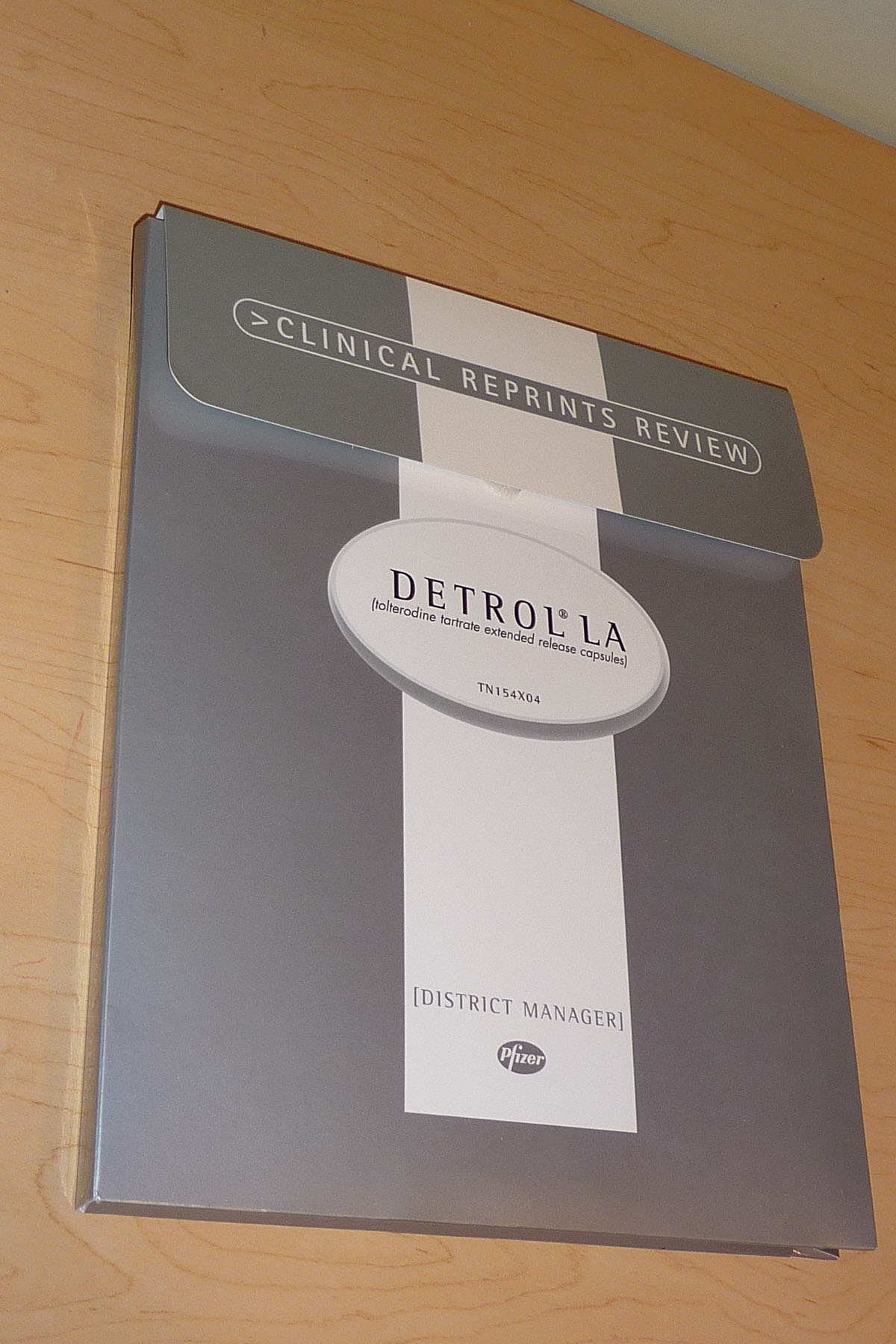 A photograph of a folder with the title Clinical reprints review : Detrol LA (tolterodine tartrate extended release capsules TV15t4X04) for District Managers at Pfizer. The book is lying on a wooden surface. The cover of the book is grey and white with text in white black and gray. The title is written in a bold font and is centered on the cover.