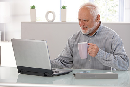 An elderly man sitting at a desk with a laptop in front of him. He is holding a pink coffee mug in his right hand and appears to be enjoying a cup of coffee. The man has a white beard and is wearing a blue checkered shirt. He has a smile on his face and seems to be deep in thought. The desk is made of glass and there are two potted plants on the windowsill behind him. The background is blurred but it seems like he is in an office or a workspace.