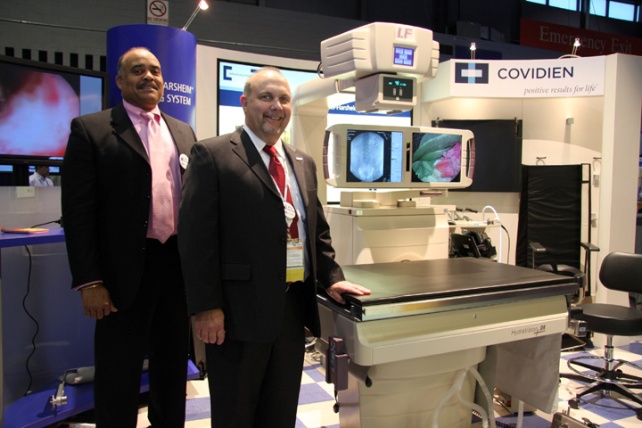 Two men standing in front of a medical equipment display at an exhibition. The display is called "COVIDIEN" and it appears to be a CT scan machine. The men are wearing suits and are smiling at the camera. Behind them there is a large screen displaying an image of a person's face. On the right side of the image there are several other medical equipment including a chair and a table. The background shows a blue and white wall with a sign that reads "Emergency Exit".