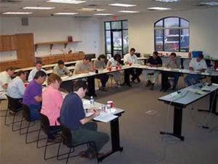 A large room with a long table in the center. The room has a high ceiling with recessed lighting and large windows on the right side. There are several people sitting at the table facing the front of the room and engaged in a discussion. They are all wearing casual clothes and appear to be of different ages and genders. On the table in front of them there are papers cups and other materials scattered across the table. There is a desk on the left side of the image with a computer monitor and other office supplies on it. The walls are painted white and there is a wooden shelf on the far wall.