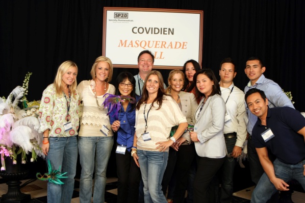 A group of people posing for a photo in front of a sign that reads "Covidien Masquerade Ball". There are nine people in the photo nine women and one man all of whom are smiling and looking at the camera. The group is standing on a stage with a black curtain in the background. On the left side of the image there is a large vase with pink and white feathers. The people are dressed in formal attire with some wearing name tags.