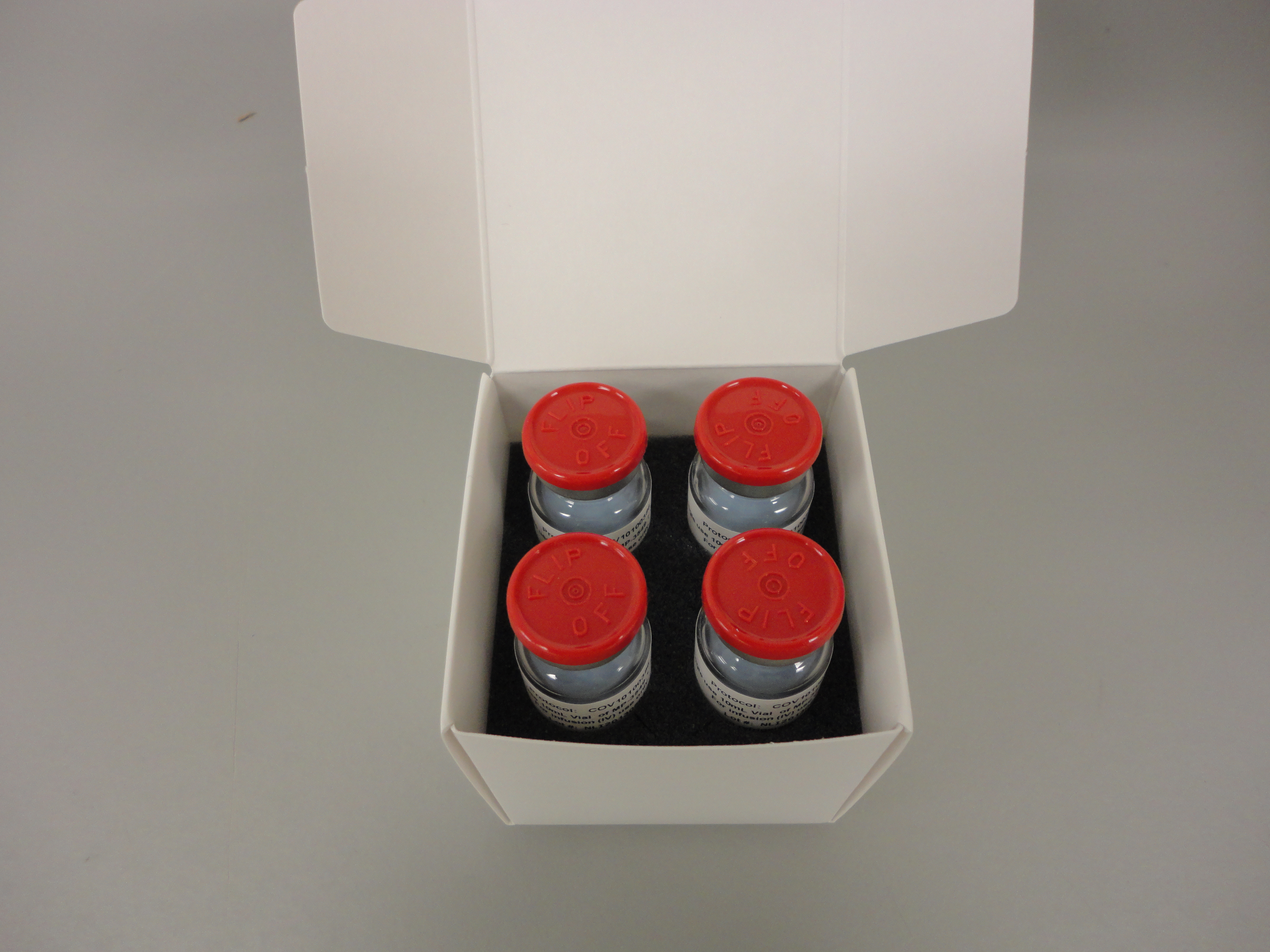 A white cardboard box with four small jars inside. The jars are arranged in a row and each jar has a red lid. The lid is slightly open revealing the contents inside the box. The box appears to be empty and is placed on a plain white surface.