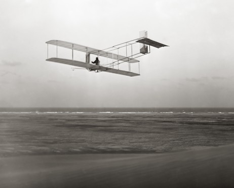 A black and white photograph of an old biplane flying over the ocean. The biplane is in the center of the image with the ocean below and the sky above. The ocean is vast and stretches out to the horizon with small waves crashing onto the shore. The sky is overcast and the horizon line is visible in the distance. The photograph appears to be taken from a low angle looking up at the biplane as it soars through the air.