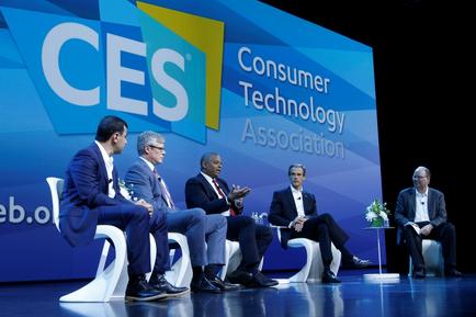 A panel discussion at the Consumer Technology Association (CES) conference. There are six men sitting on a stage in front of a large blue screen with the CES logo and the words "Consumer Technology Association" written in white. The men are dressed in formal business attire and appear to be engaged in a discussion. They are sitting on white chairs and are facing each other with one man gesturing with his hands as he speaks. On the right side of the stage there is a small table with a vase of flowers. The background is dark suggesting that the conference is taking place at night.