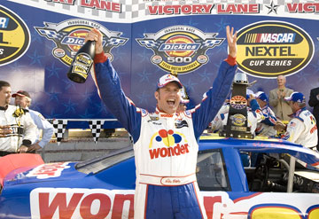 A man standing on top of a race car with his arms raised in triumph. He is wearing a blue and white racing suit with the word "Wonder" written across the chest. He has a big smile on his face and is holding up a bottle of champagne in his right hand. In the background there are several other race cars and a banner that reads "Victory Lane" and "Nasar Nextel Cup Series". There are also several people standing around the car cheering and celebrating. The image appears to have been taken at a NASCAR race track.