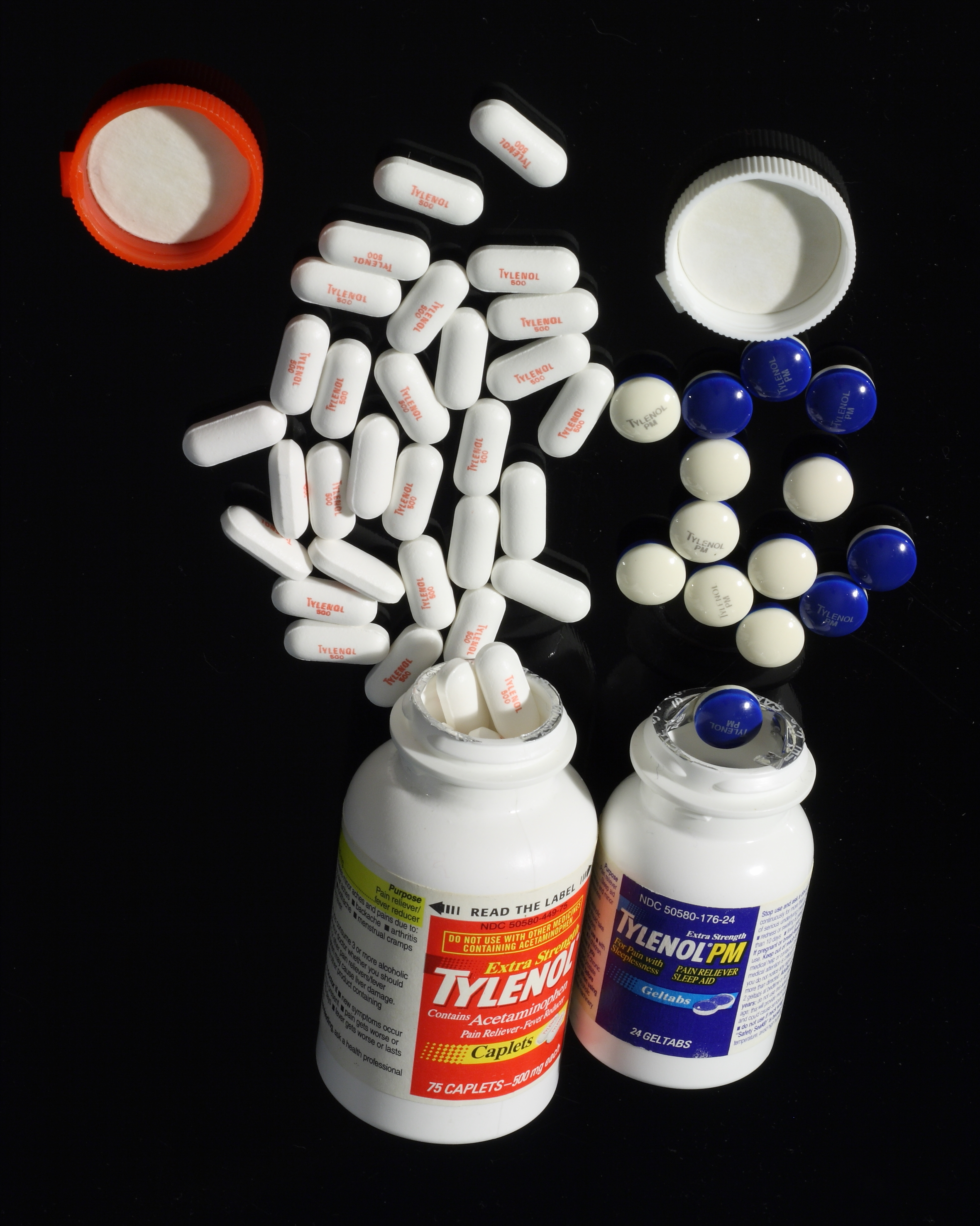 Two white bottles with blue caps lying on a black surface. The bottles are labeled "Tylenol" and "Tylenol PM". There are several white pills scattered around the bottles some of which are spilling out of the caps. The pills are of different sizes and colors including red white and blue. There is also an orange pill bottle cap on the top left corner of the image. The background is also black making the colors of the pills stand out.