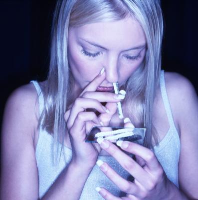 A young woman with blonde hair wearing a white tank top holding a pack of cigarettes in her hands. She is looking down at the pack with a sad expression on her face. The background is dark and the lighting is blue creating a moody atmosphere. The woman appears to be deep in thought with her eyes closed and her mouth slightly open.