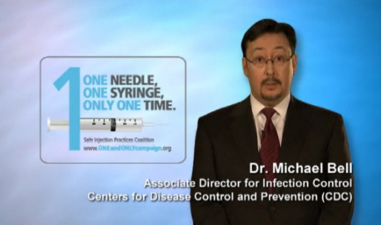 A screenshot from a video interview with Dr. Michael Bell Associate Director for Infection Control Centers for Disease Control and Prevention (CDC). He is wearing a suit and tie and is standing in front of a blue background. On the left side of the image there is a logo for One Needle One Syringe Only One Time. The logo is a syringe with the text "One Needle" and "One Syringe" above it. Below the logo there are two lines of text that read "Safe Injection Practices Coalition" and the website "www.oneandonlycampaign.org".