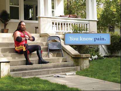 A man sitting on the steps of a house. He is wearing a red tank top black pants and black boots. He has long dark hair and a beard. He appears to be deep in thought with a serious expression on his face. There is a blue sign next to him that reads "You know pain." The house has white columns and a porch with a railing. There are trees and bushes in the background.