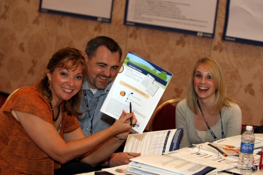 Three people sitting at a table in a conference room all of whom are smiling and looking at a brochure. The man is holding the brochure in his hand and appears to be explaining it to the two women on either side of him. The woman on the left is wearing an orange blouse and the woman in the center is wearing a gray blouse. There are several other brochures and papers on the table in front of them. In the background there are two whiteboards and a projector screen.