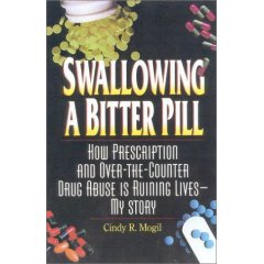 The cover of a book titled "Swallowing a Bitter Pill: How Prescription and Over-the-Counter Drug Abuse is Ruining Lives-My Story" by Cindy R. Mogil. The cover is predominantly black with white text. The title of the book is written in large bold font at the top of the cover. Below the title there is a small illustration of a jar of pills spilling out of it. The jar is filled with yellow pills and there are several other pills scattered around the jar. The background is a dark blue color. The author's name and the publisher's name are written in smaller white text at the bottom.