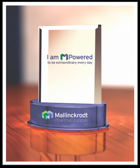 A digital illustration of a trophy or award. The trophy is rectangular in shape and has a blue base with the logo of Mallinckrodt Pharmaceuticals on it. The logo is a green and blue shield with the company's name written in white text. The background is blurred but it appears to be a wooden table or desk. The text on the trophy reads "I am M Powered to be extraordinary every day". The overall design of the trophy is modern and professional.