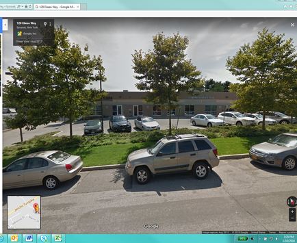 A street view of a parking lot with several cars parked in it. The parking lot is located in front of a building with a brown exterior and a white roof. There are several trees and bushes surrounding the lot and a few cars are parked in the lot. The sky is cloudy and the overall mood of the image is gloomy.
