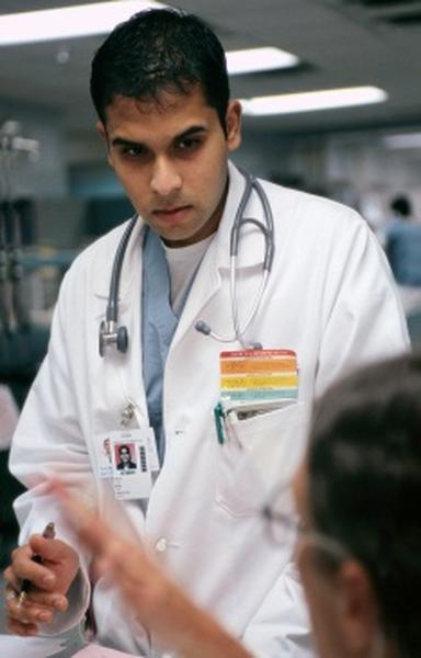 A young man in a white lab coat with a stethoscope around his neck. He is sitting at a desk and appears to be engaged in a conversation with another person. The man is holding a pen and seems to be explaining something to the other person who is sitting in front of him. The background is blurred but it seems like the focus is on the two people in the foreground. It seems like they are in a hospital or medical facility.