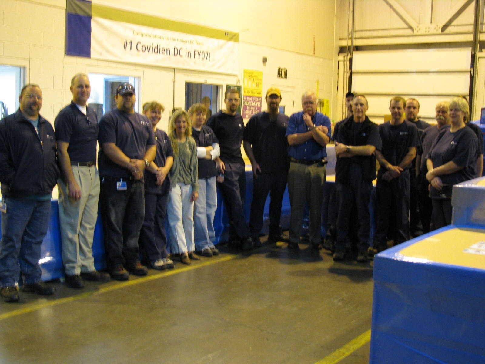 A group of 16 people standing in a warehouse posing for a group photo. Most of them are wearing dark shirts and work pants. They are standing in front of a banner that says "Congratulations to the [illegible] DC Team : #1 Covidien DC in FY07!" A dock door pallets of boxes and windows (in offices?) are visible in the background.