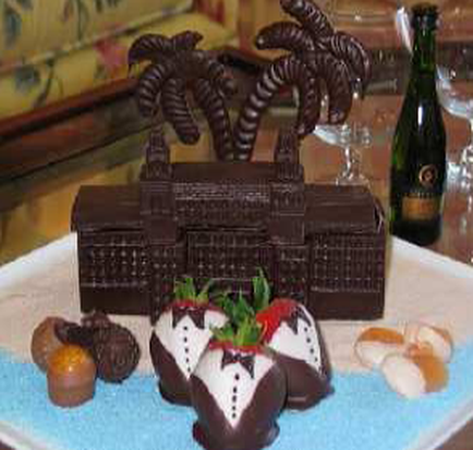 A chocolate cake on a blue plate. The cake is in the shape of a castle with two palm trees on top. The castle is made of dark chocolate and has a pointed roof. On the plate there are three chocolate strawberries with white frosting and green leaves on top arranged in a pyramid-like shape. There are also a few chocolate truffles scattered around the plate. In the background there is a bottle of champagne and a glass of wine on a table.