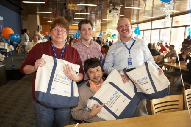 Four people two men and two women standing in a large room with tables and chairs. They are all smiling and holding large blue and white bags with the word "EXALGO" written on them. The woman on the left is wearing a red sweater and glasses the man in the middle is wearing an orange shirt and the man on the right is wearing glasses and a blue lanyard. The room is decorated with blue and orange balloons and there are other people in the background. It appears to be an event or gathering.