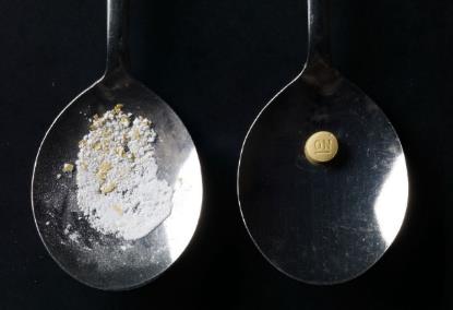 Two silver spoons side by side on a black background. The spoon on the left is filled with a white powdery substance which appears to be a mixture of herbs and spices. On the right spoon there is a small gold-colored coin with the word "ON" engraved on it. The coins are placed on the right side of the spoon. The overall mood of the image is dark and mysterious.