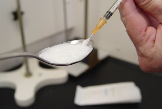 A hand holding a syringe inserted into a spoon full of white liquid. The spoon is above a black countertop with various small items nearby. The background is blurred but it appears to be a laboratory setting.