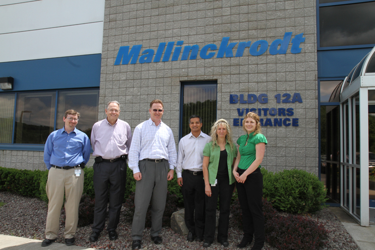 A group of six people standing in front of a building with a blue sign that reads "Mallinckrodt Bldg 12A Visitors Entrance". The building appears to be a modern office building with large windows and a glass door. The group is standing in a line facing the camera and smiling. There are four men and two women all in business casual clothing. They are standing on a bed of gravel with bushes and shrubs behind them.