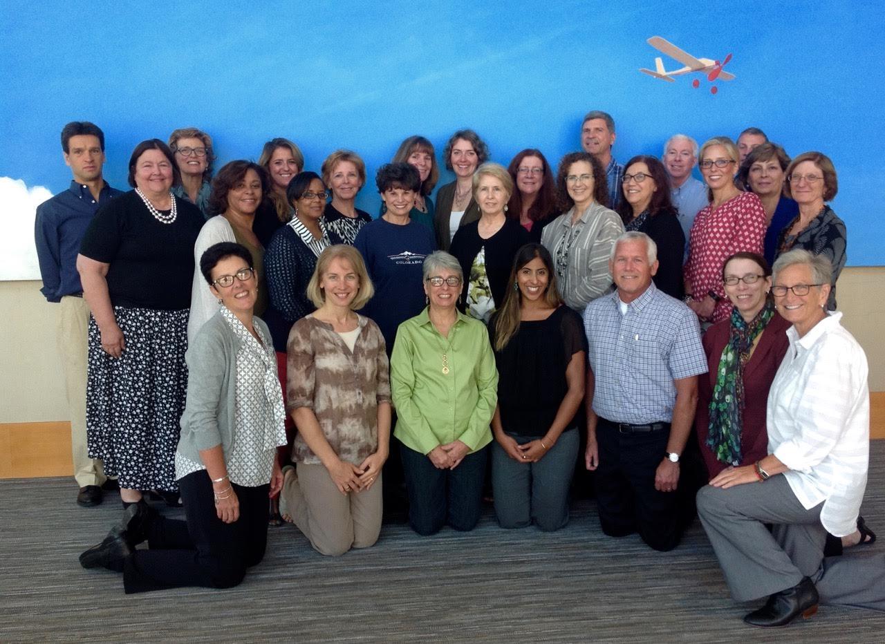 A group of people posing for a group photo in front of a blue wall with an airplane painted on it. There are around 20 people in the photo all of them are smiling and looking at the camera. Some are standing and some are kneeling. The group appears to be of different ages and ethnicities and they are all dressed in casual clothing. The airplane is painted in red and white and it is flying in the sky. The background of the photo is a gray carpeted floor.