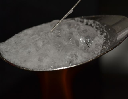 A close-up of a spoon containing a bubbly white substance with a needle inserted into the substance. The spoon is gray and the background is solid black.