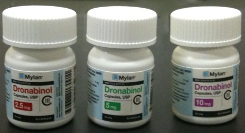 Three white plastic bottles with white caps. Each bottle has a blue label with the brand name "Mylan" and the product name "Dronabinol" printed on it. The bottles are arranged in a row with the first bottle on the left the second bottle in the middle and the third bottle at the bottom. All three bottles appear to be new and unused. The labels also mention that the bottles contain 2.5 mg of Dronabinol capsules USP and 10 mg of the product. The background is a plain black surface.