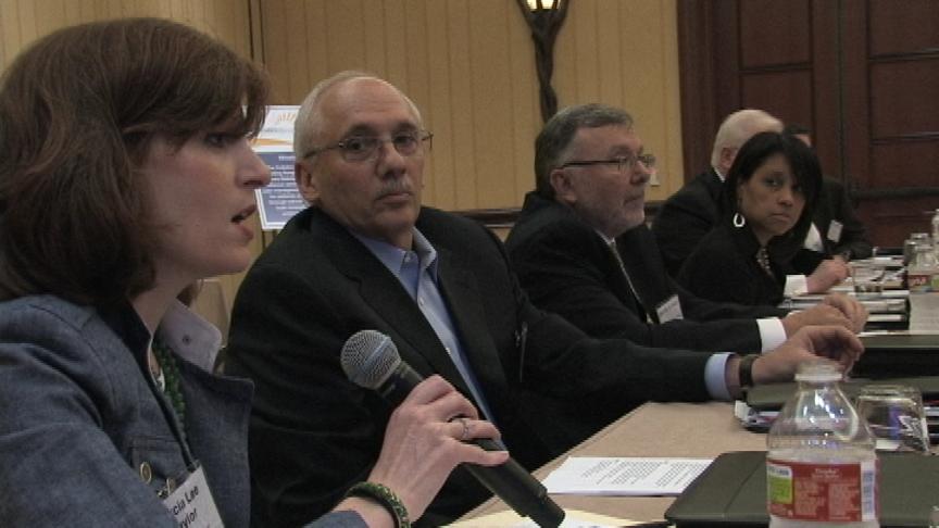 A group of people sitting at a long table in a conference room. There are four people in the image three men and two women all of whom appear to be engaged in a discussion. The woman on the left is holding a microphone and appears to be speaking while the man in the middle is listening attentively. The man on the right is wearing a suit and glasses and the woman next to him is also speaking into the microphone. The table is covered with papers and water bottles and there is a projector screen in the background. The room has wooden paneling and a chandelier hanging from the ceiling.