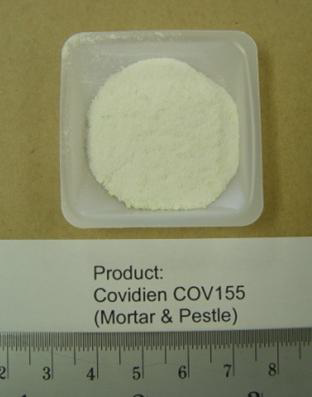 A small square plastic container with a white powder inside. It is labeled as Covidien COV155 (Mortar & Pestle). The container is placed on a beige surface with a ruler at the bottom for scale.