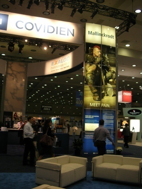 A trade show booth with a large banner that reads "Covidien" and "Mallinckrodt" on the right side. On the left side of the booth there is a sign that says "Exalgo" and on the bottom right corner there are two white sofas. In the center of the image there appears to be a large poster with an image of a man holding a gun and the text "Meet Pain". There are several people in the background some of whom are walking around the booth. The booth is brightly lit with spotlights and there are other booths and signs visible.