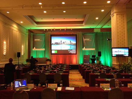 A large conference room with rows of tables and chairs set up for a meeting. The room has a high ceiling with recessed lighting and a chandelier hanging from it. The walls are covered in green curtains and there is a large screen in the center of the room displaying a presentation. On either side of the screen there are two large screens displaying a city skyline. The tables are covered with red tablecloths and have nameplates water bottles and laptops on them. There are also a few people in the room suggesting that the meeting is taking place in a formal setting.