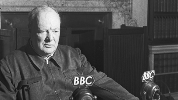 A black and white photograph of an elderly man sitting at a desk with two microphones in front of him. The man is wearing a jacket and appears to be in his late 60s or early 70s. He has a serious expression on his face and is looking off to the side. The microphones have the letters "BBC" on them indicating that they are from the British Broadcasting Corporation (BBC). The background is a room with a fireplace and a bookshelf.