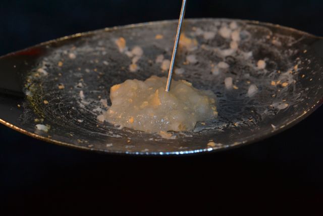 A frying pan with a small amount of food in it. The pan appears to be made of stainless steel and has a shiny surface. The food in the pan is a light yellow color and looks like it is being cooked. A metal spatula is being used to stir the food. The background is dark and out of focus making the frying pan the focal point of the image.