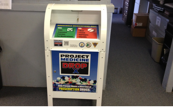 A white vending machine with a blue and red poster on it. The poster has the words "Project Medicine Drop" written in bold letters at the top followed by "Prescription Drugs" in smaller letters. Below the poster there is a list of prescription drugs and their corresponding prices. The vending machine is located in an office or storage area with a gray carpeted floor and a white wall in the background.