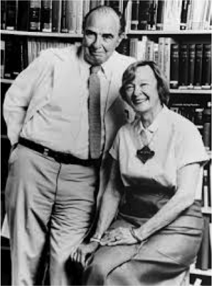 A black and white photograph of an elderly couple posing for a photo in front of a bookshelf. The man is on the left side of the image wearing a suit and tie and the woman is sitting on the right side. They are both smiling and looking at the camera. The woman is wearing a dress with a badge on her lapel and has her hands clasped together. The background is filled with bookshelves suggesting that the photo was taken in a library or study. The couple appears to be happy and relaxed with the man's arm around the woman's waist.