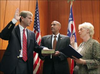 Three people standing in a room with wooden walls and two American flags in the background. On the left side of the image there is a man in a suit and tie who appears to be giving a high-five to a woman in a military uniform. The man in the middle is holding a book and the woman on the right side is clapping her hands in appreciation. It appears that the man is receiving an award or recognition from the woman.