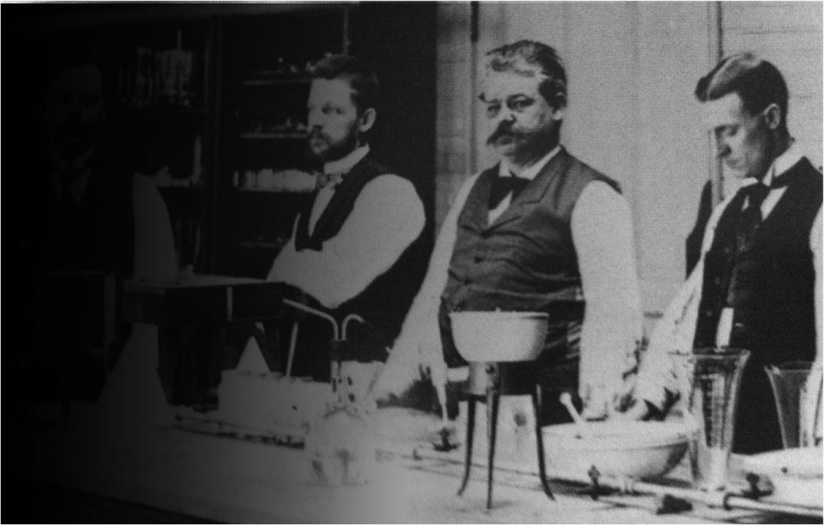 A black and white photograph of three men in a laboratory setting. The men are standing in front of a table with various scientific equipment and equipment on it. The man in the center is wearing a vest and has a mustache and appears to be in his late twenties or early thirties. He has a serious expression on his face and is looking off to the side. On the left side of the image there is another man with a beard and a mustache who is sitting at a desk with a microscope and other scientific equipment. The other two men are wearing suits and appear to be observing the experiment. The background is blurred but it seems like the men are focused on their work.