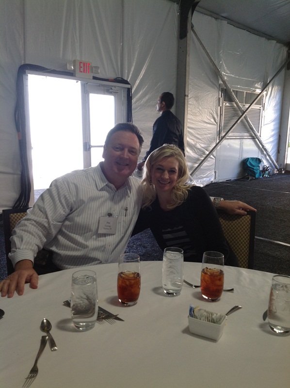 A man and a woman sitting at a table in a tent. They are both smiling and appear to be happy. The table is covered with a white tablecloth and there are several glasses of drinks on it. The man is wearing a striped shirt and has a name tag around his neck. The woman is also smiling and has blonde hair. In the background there is another person standing near a window and an exit sign. The tent appears to be set up for a party or event.